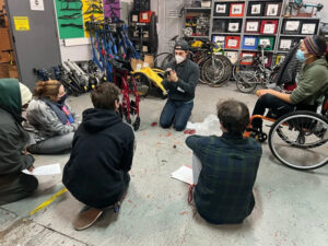 A group of individuals listen and watch a speaker helping them to understand the process of making repairs to a wheelchair. They are young adults of varying ages and ethnicities. Four of the five listeners and the speaker are seated while the fifth sits in a wheelchair. Beyond them are wheelchair parts, bikes, and bike repair parts.