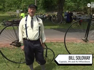 Bill Soloway is interviewed by 6ABC as the caption of the NJ and PA chapters of the Wheelmen with two big-wheel antique bicycles behind him in a public park. Bill wears old-fashioned dress with a buttoned shirt, a tie, suspenders, and black pants to the knee. He has light skin, gray hair, and glasses.