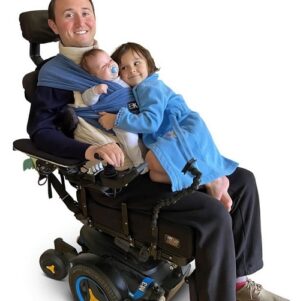Quadriplegic father Josh Basile is pictured with his infant son and young daughter. His son is strapped to his chest with a sling and his daughter is wearing a blue bath robe and cuddling her younger brother as she sits on Josh's lap. Josh has light skin, short dark hair, and a big smile as he sits in his black power chair.