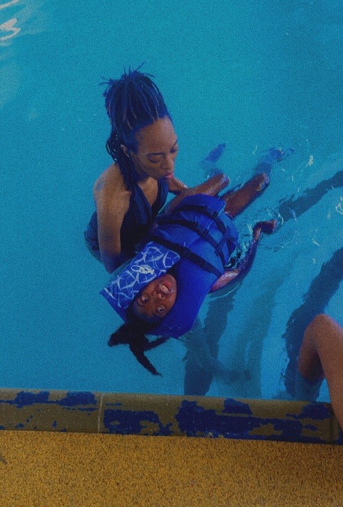Mom Sierra holds Zoë afloat in a pool. Sierra has brown skin, braided black hair, and a navy swimsuit. Zoë has brown skin, black textured hair, and a blue life vest that covers most of her body and torso from the neck down.