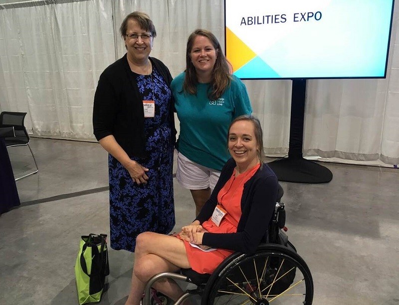 At an Abilities Expo convention, Help Hope Live's Sonny Mullen wears a teal t-shirt with the Help Hope Live logo and shorts as she smiles for a photo with Cindy Kolbe, who is wearing a blue and black pantsuit and has light skin and short gray-brown hair and glasses. They are with Cindy's daughter, Beth, who has light skin, brown hair, gold hoop earrings, and a pink dress as she sits in her black manual wheelchair.