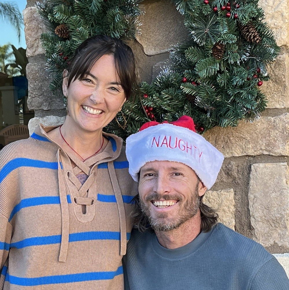 Megan and Arthur are in front of a stone wall with a holiday wreath. Megan has dark hair and light skin and wears a striped hoodie. Arthur has light skin, a short beard, and brown hair to his shoulders and wears a Santa hat that says NAUGHTY.