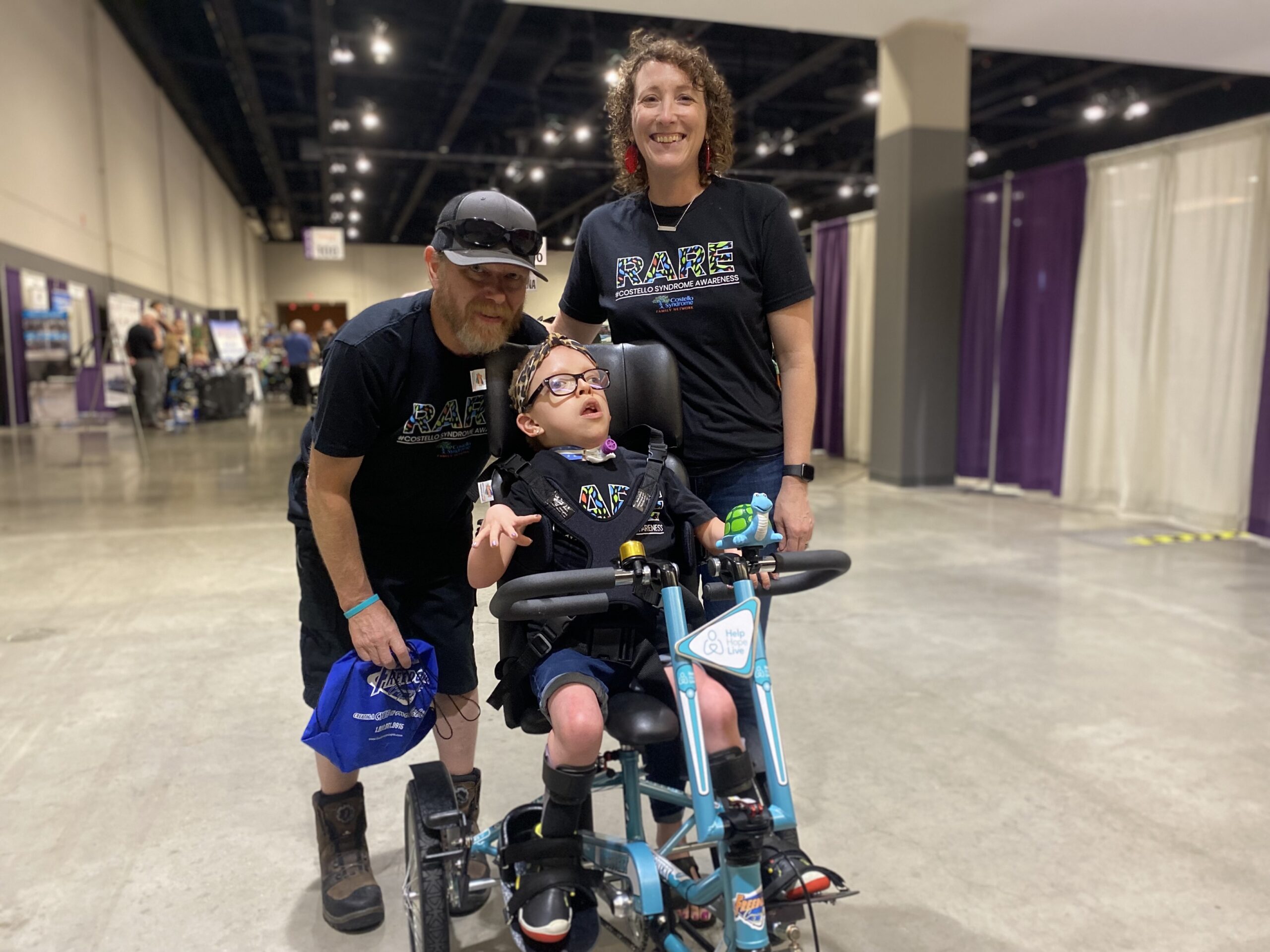 Anna-Stella Roberts is an 11-year-old with light skin and glasses who is sitting on her new adaptive bike in Help Hope Live teal. Her parents stand behind her with matching t-shirts that read RARE: Costello Syndrome Awareness. They are at Abilities Expo Chicago 2023.