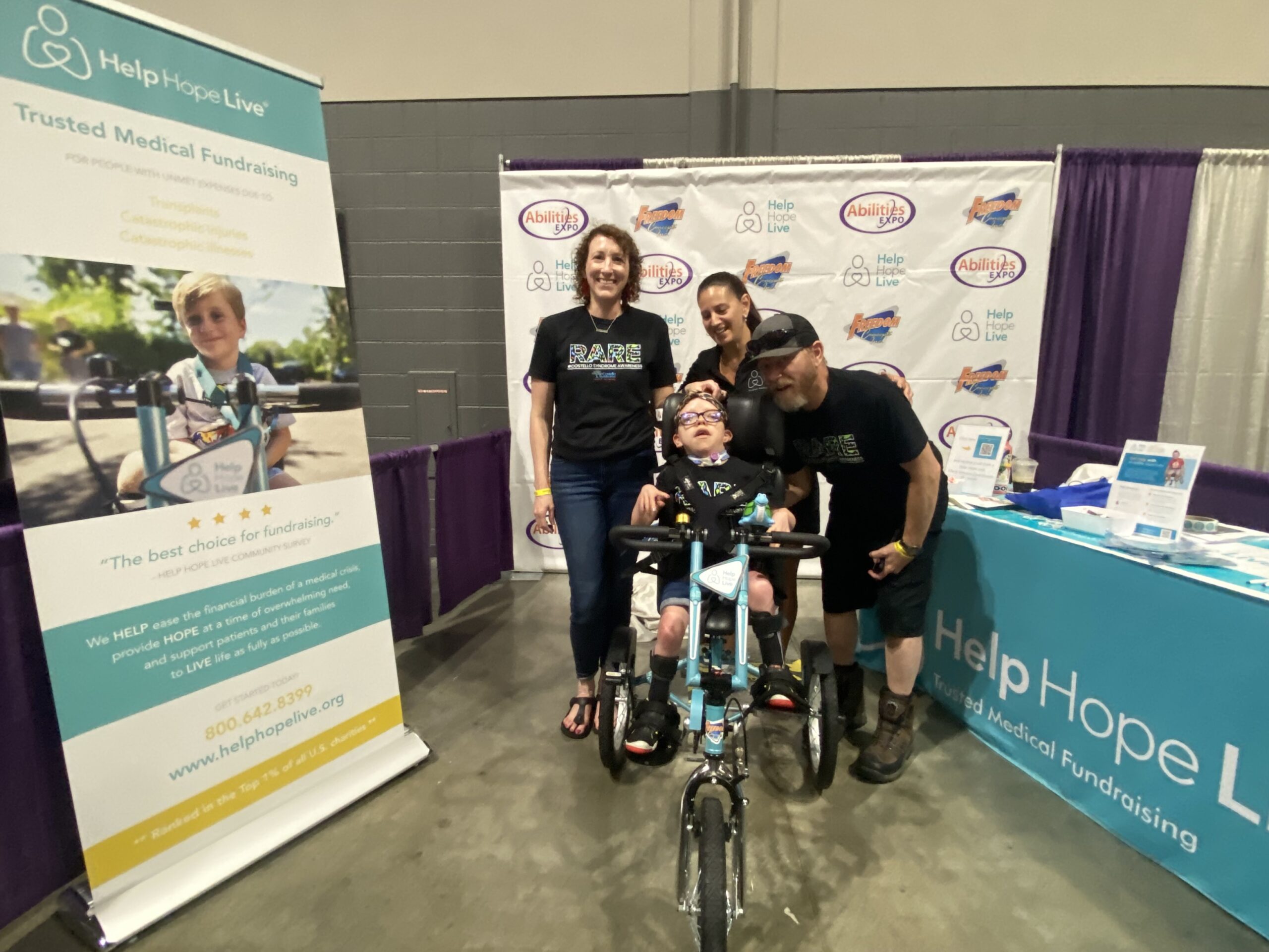 At the Help Hope Live booth of Abilities Expo Chicago, Anna-Stella's parents stand next to her and smile along with Help Hope Live's Kelly L Green as Anna-Stella sits on her new Help Hope Live themed adaptive bike. Anna-Stella has light skin and glasses.