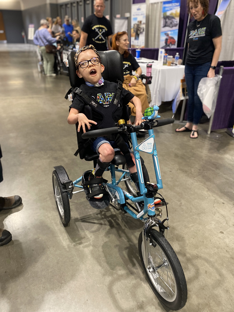 11-year-old Anna-Stella Roberts is seated on her new teal Help Hope Live themed adaptive tricycle from Freedom Concepts at Abilities Expo Chicago 2023. She has light skin, brown eyes, and glasses.