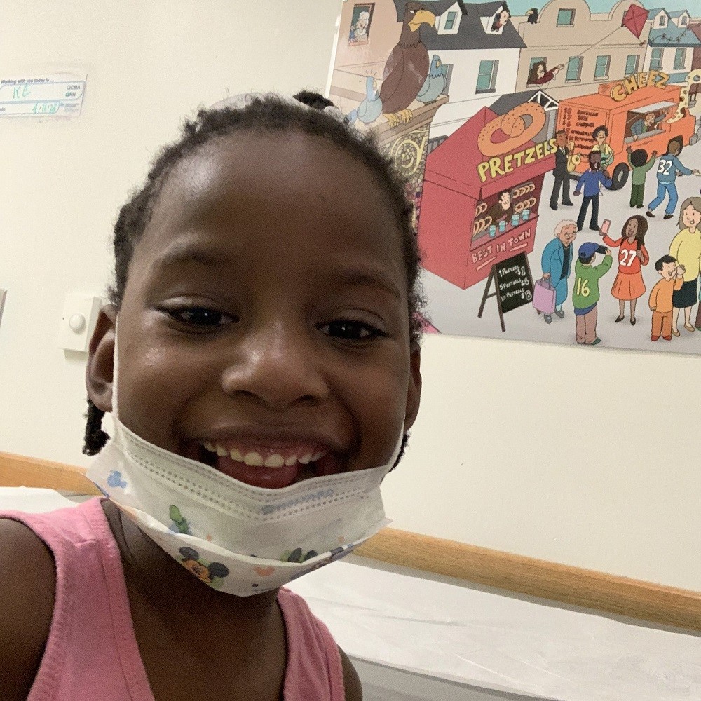 Divinity McFarland is a little girl with brown skin and short black braided hair. She is smiling with a mask over her chin. Behind her is a colorful cartoon city scene.