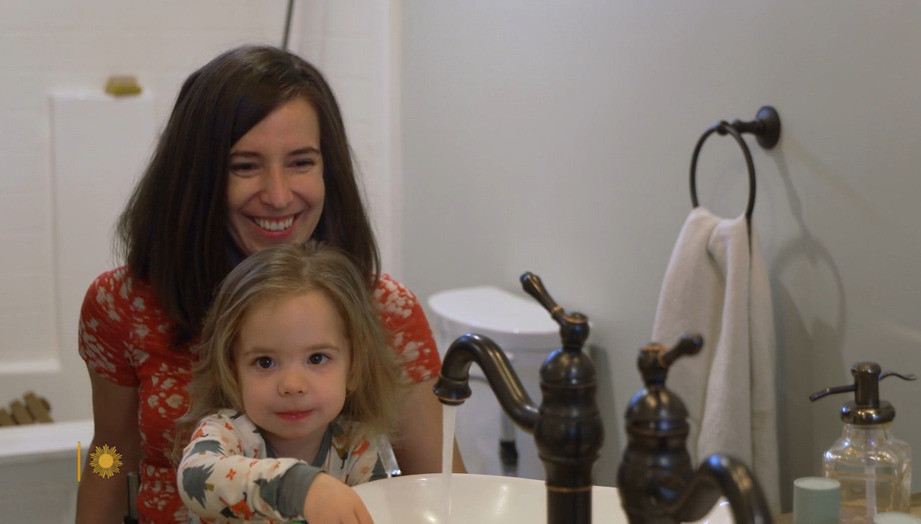 Quadriplegic mom Daniela Izzie with one of her twin daughters who is brushing her teeth at the sink. Dani has light skin and brown straight hair past her shoulders. Her daughter has light skin, brown eyes, and blonde hair.