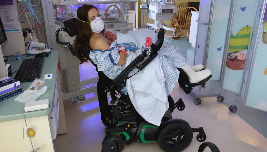 In the hospital after giving birth, Daniela Izzie is seated in her black power chair with a hospital gown and mask holding her newborn twin daughters. Dani has light skin and straight brown hair.