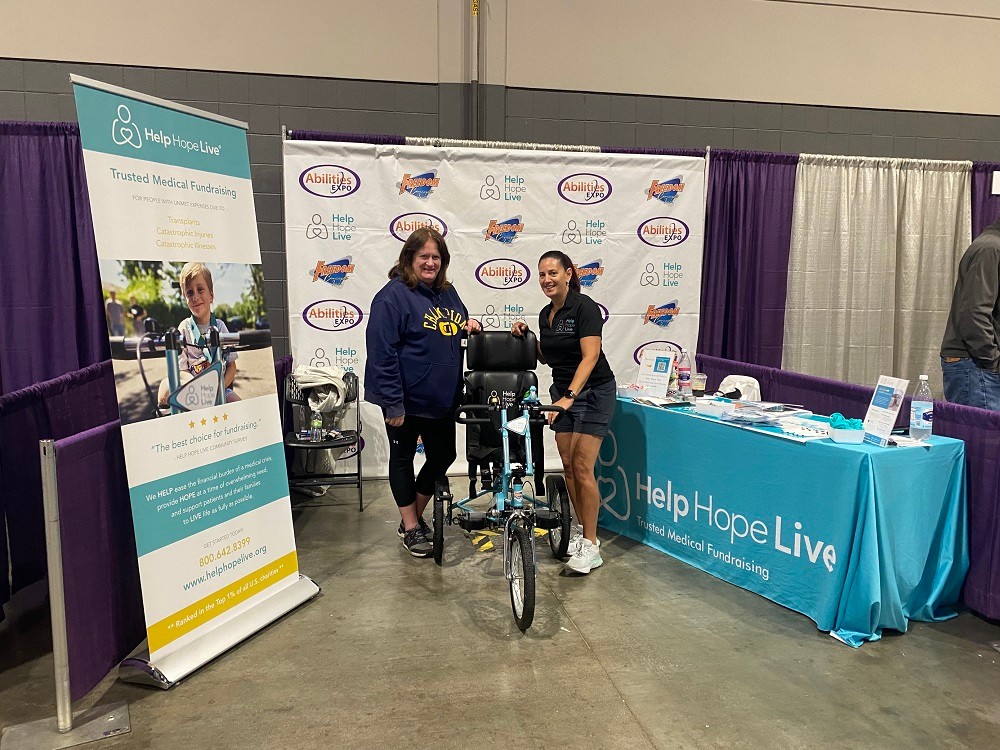 The Bike Rack's Tammy Roberts stands next to Help Hope Live's Kelly L Green at the Help Hope Live booth of Abilities Expo Chicago - between them is a new adaptive bike with Help Hope Live details and teal coloring.