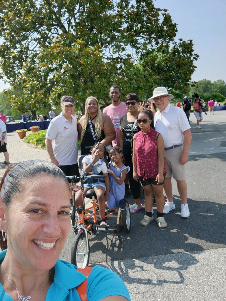 Help Hope Live Executive Director Kelly L Green takes a selfie with Divinity McFarland visible behind her on her new adaptive bike in Flyers orange. With brown skin and braided dark hair, Divinity is with legal guardian Elsie, who has brown skin and long blonde braids, other family members, and two members of the Flyers Alumni Association, both white men in golf attire.