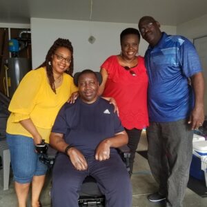 Stephany is a male with brown skin and brown eyes seated in his power chair. His wife Katrina stands beside him with brown skin, textured brown hair, and glasses along with two friends.