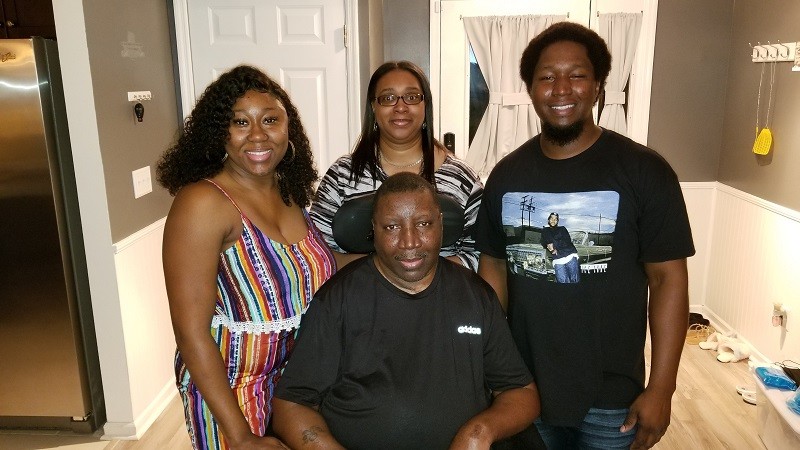 Stephany is a man with brown skin and brown eyes seated in a black power chair. Behind him is his wife Katrina, who has brown skin, glasses, brown eyes, and straight brown hair, and two younger people. They are in the entrance hallway of a home.