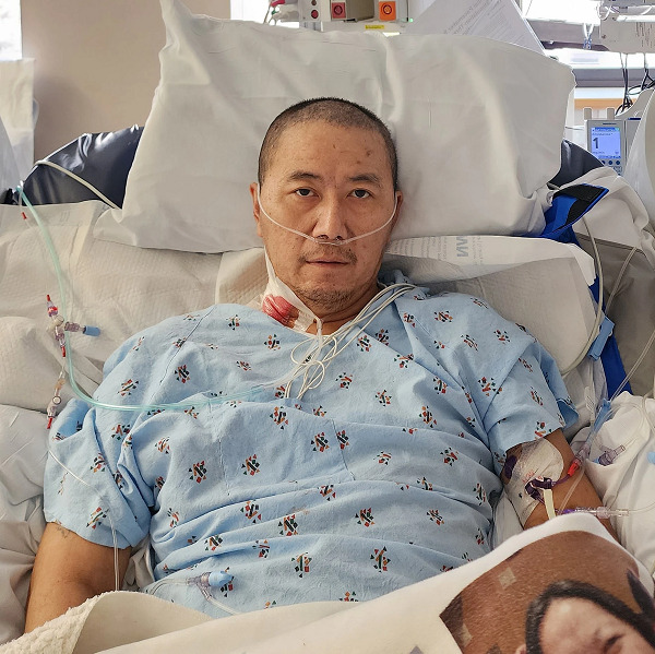 Kue is an Asian man with a shaved head and dark eyes. He is in a hospital bed wearing a hospital gown with an oxygen tube connected to his nose.