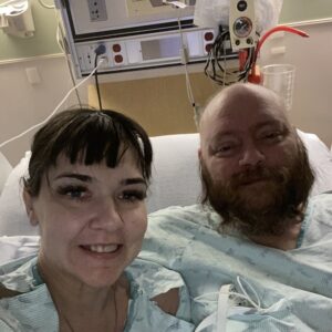 Michelle and her husband and kidney donor are lying in a hospital bed, both wearing hospital gowns. Michelle has light skin, dark eye makeup, and dark hair with short bangs. Her husband has light skin, a bald head, and a big red beard.