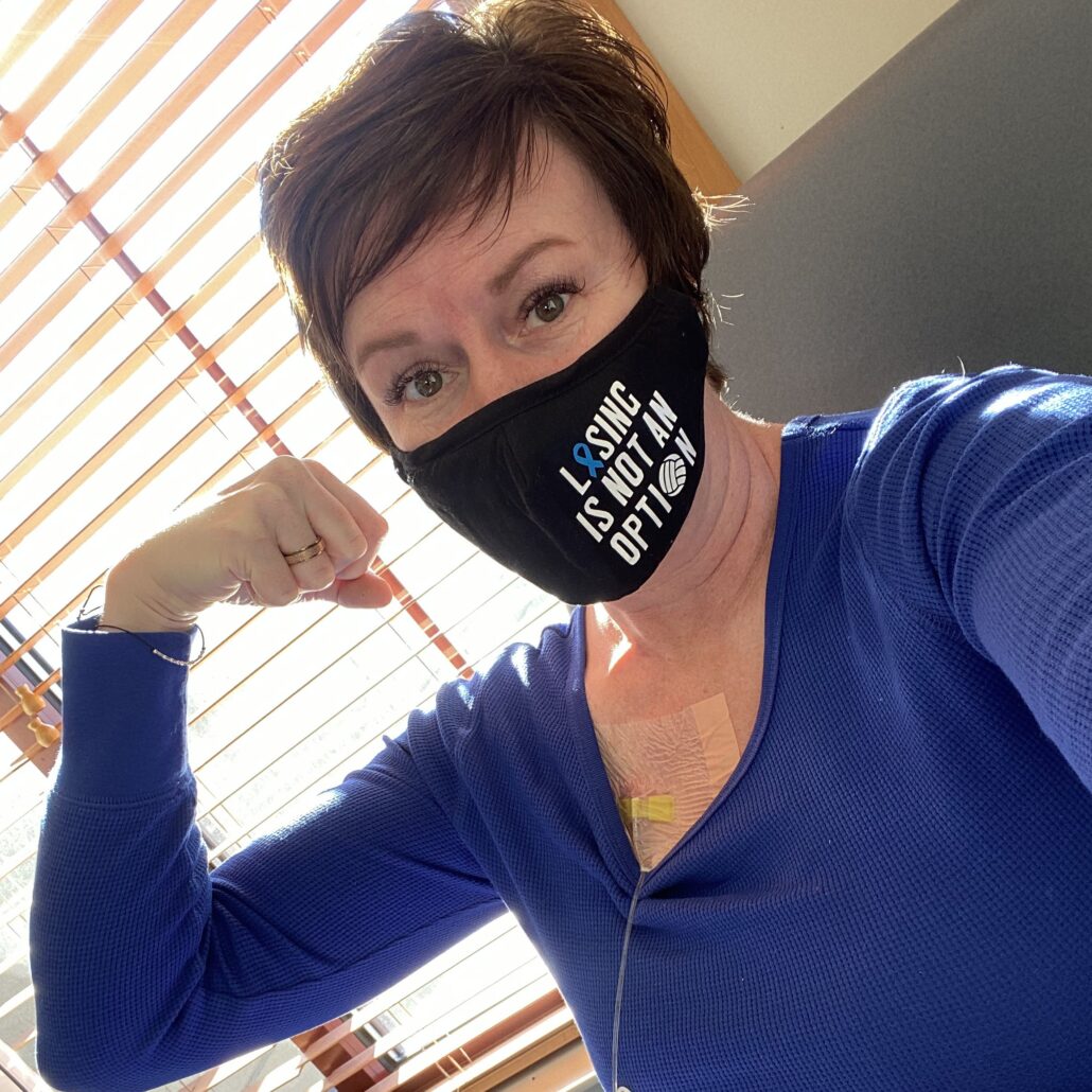 Natalie Jo Stika has light skin, short brown hair, and brown eyes and wears a black face mask that reads Losing is Not an Option. She wears a purple long-sleeved shirt and is making a muscle with her right arm.