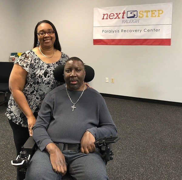 Stephany is a man with brown eyes and brown skin seated in his power chair. He wears a cross chain. His wife Katrina stands behind him. She has brown skin, glasses, brown eyes, and straight dark hair. They are at Next Step Raleigh Paralysis Recovery Center.