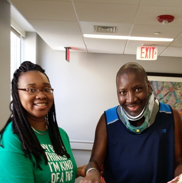 Stephany and Katrina Golden both smile as Katrina holds Stephany's hand as he stands with assistance. Stephany has brown skin and brown eyes and wears a neck brace. Katrina has brown skin, brown eyes, glasses, and textured black hair in braids.