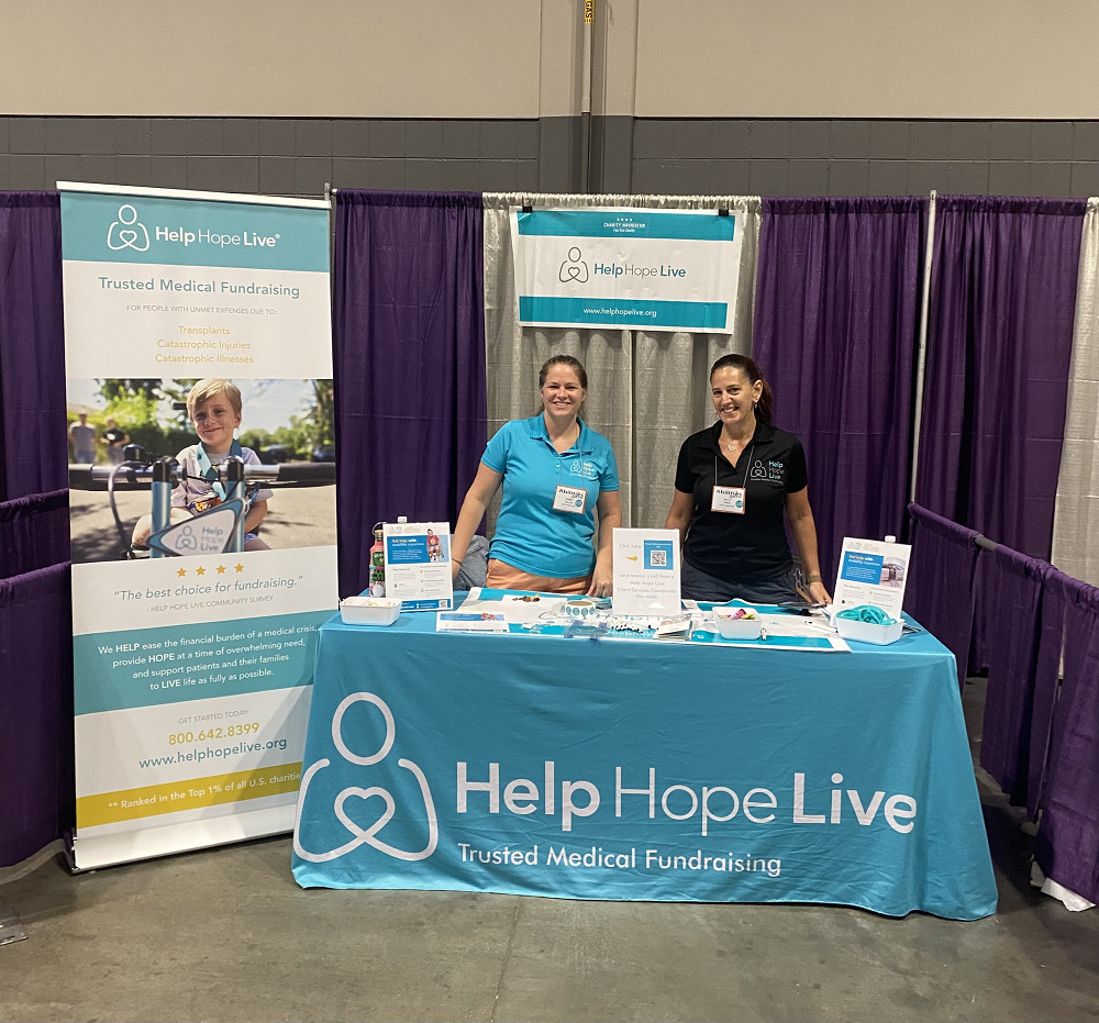 Kelly L Green and Sonny Mullen are at the Help Hope Live booth of Abilities Expo Chicago 2023. The table features a teal Help Hope Live: Trusted Medical Fundraising tablecloth with materials, pens, bracelets, and more. Sonny has light skin and sandy hair pulled back and wears a teal Help Hope Live polo shirt. Kelly L Green has light skin and dark hair pulled back with a black Help Hope Live polo shirt.