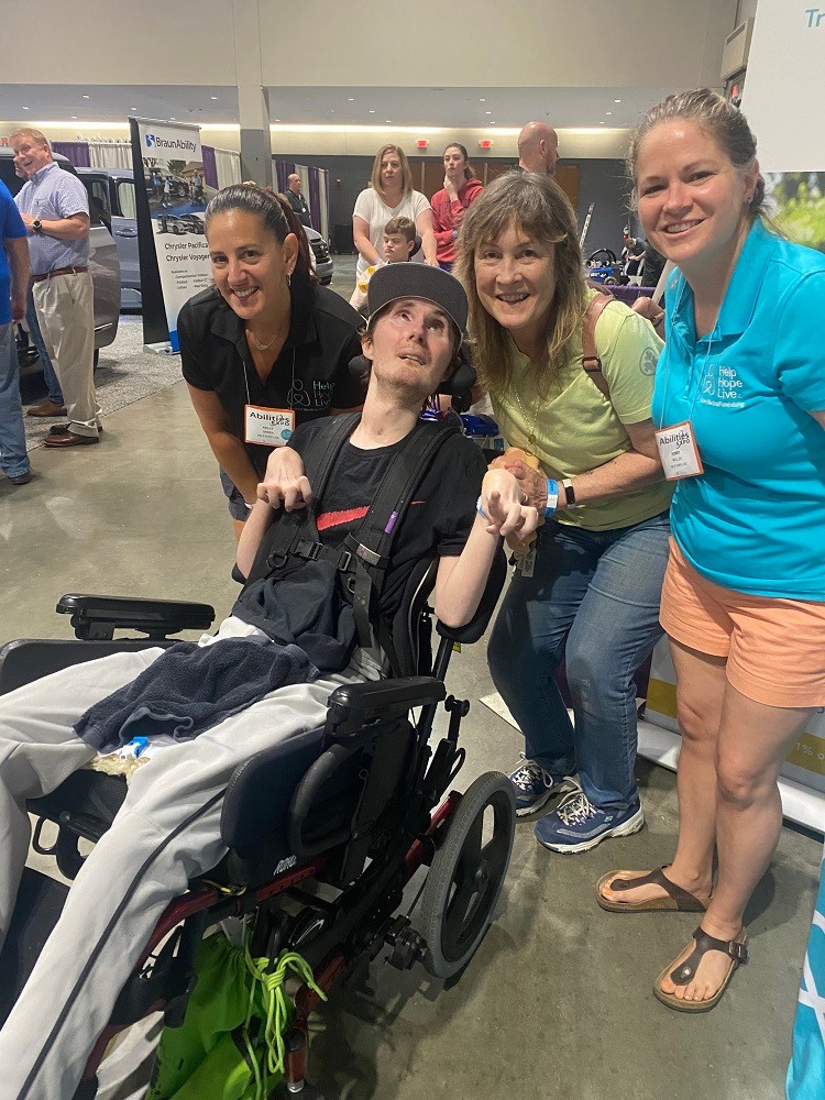 Help Hope Live's Kelly L Green and Sonny Mullen meet client Nick Nigro and his mom Lisa Nigro at Abilities Expo Chicago. Nick has light skin, a baseball cap, and eyes looking upward as he sits in a black power chair. He wears athletic gear and his hands are slightly curled.