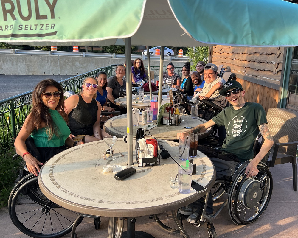 At an outdoor patio, several members of the Live to Roll team are seated in their wheelchairs ready for a meal alongside Help Hope Live's Sonny Mullen and Kelly L Green and WOW Wonders on Wheels's Justin Graham.