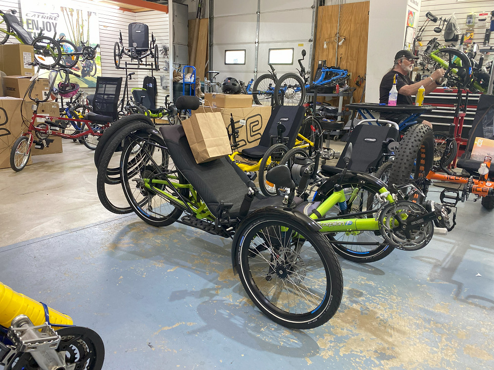 A man with gray hair and a beard works on a bike at The Bike Rack in Chicago with a seated adaptive bike visible alongside many other adaptive bikes and non-adaptive traditional bikes.