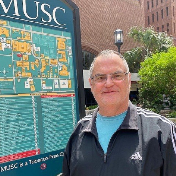 William is standing next to an MUSC campus map. He has light skin, glasses, and short gray hair.