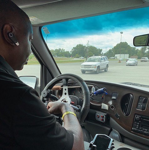 Stephany is a man with brown skin wearing an in-ear headset and driving an adapted vehicle using hand controls on the steering wheel.