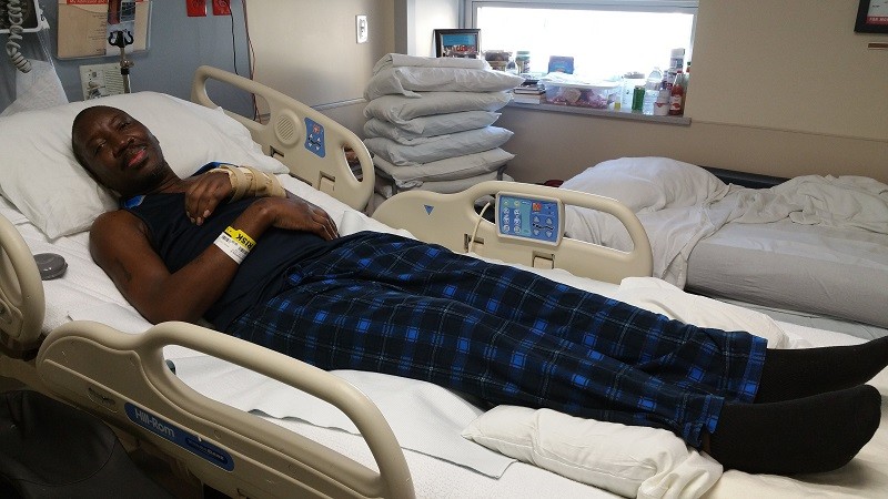 Stephany smiles as he lies in a hospital bed wearing a black tank top, black socks, and blue flannel pajama pants. He has brown skin and brown eyes and hospital bracelets.