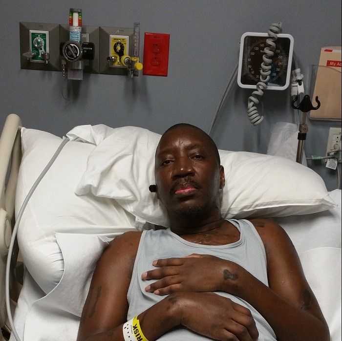 Stephany is lying in a hospital bed. He has brown skin, a short black mustache, brown eyes, and hospital bracelets.