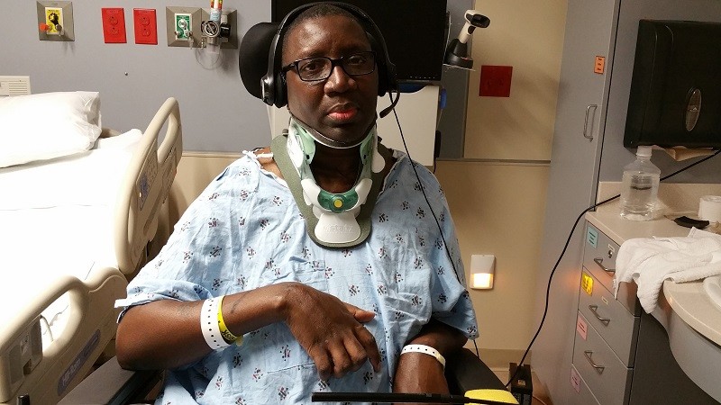 In a hospital setting, Stephany wears a hospital gown, hospital bracelets, a neck brace, and headphones. He has brown skin, brown eyes, and glasses.