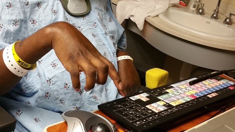 A close-up of Stephany's hands, which are slightly curled as he reaches out a finger to tap an adaptive keyboard. He is wearing hospital bracelets and a hospital gown.