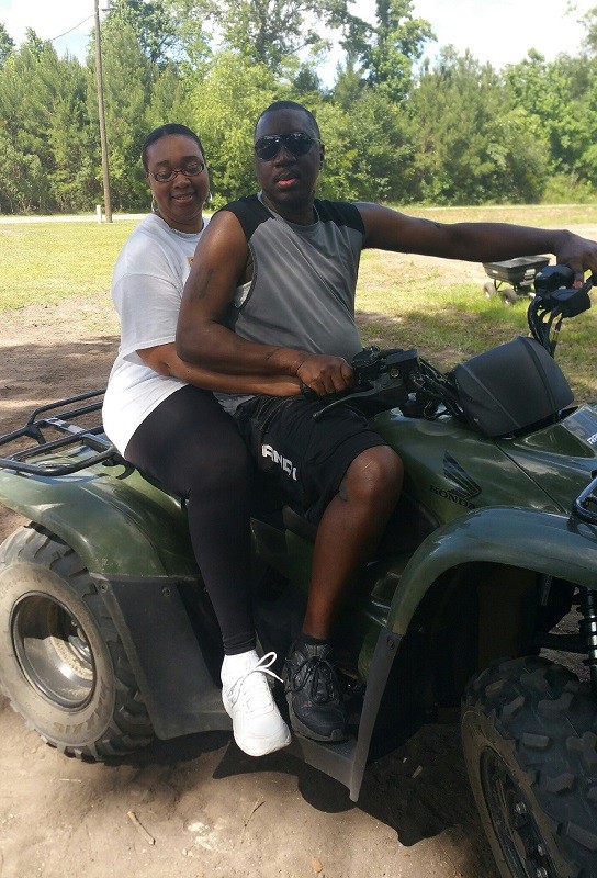 Shown before his spinal cord injury, Stephany Golden and his wife Katrina sit on an ATV outdoors. Both have brown skin and sneakers - Stephany wears sunglasses while Katrina wears glasses.