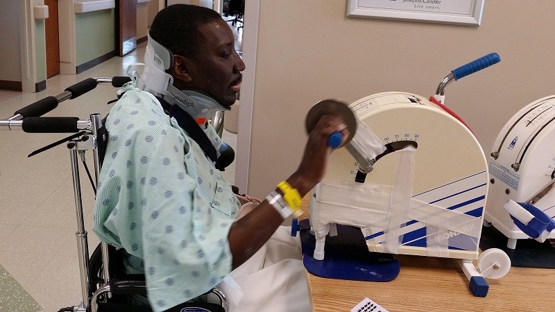 In a rehab setting wearing a hospital gown and hospital bracelets, Stephany uses a handheld exercise machine. He has brown skin and a neck brace and he sits in a manual wheelchair.