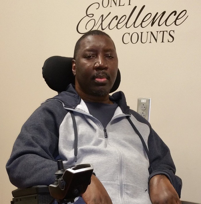 Stephany is a man with brown skin and brown eyes seated in his black power chair wearing a zip-up hoodie. Behind him is a wall decal that reads Only Excellence Counts.