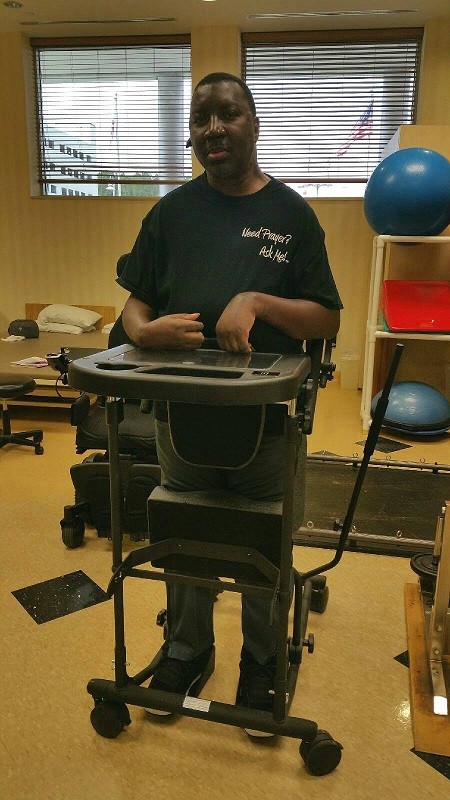 Stephany is a male with brown skin and brown eyes standing in a standing frame with a fold-out desk to rest his hands on in a therapy setting. His shirt reads Need Prayer? Ask Me!