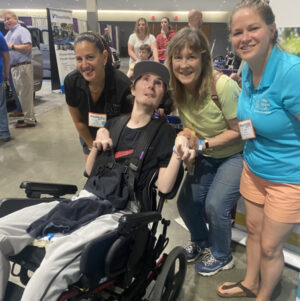 Help Hope Live's Kelly L Green and Sonny Mullen meet client Nick Nigro and his mom Lisa Nigro at Abilities Expo Chicago. Nick has light skin, a baseball cap, and eyes looking upward as he sits in a black power chair. He wears athletic gear and his hands are slightly curled.