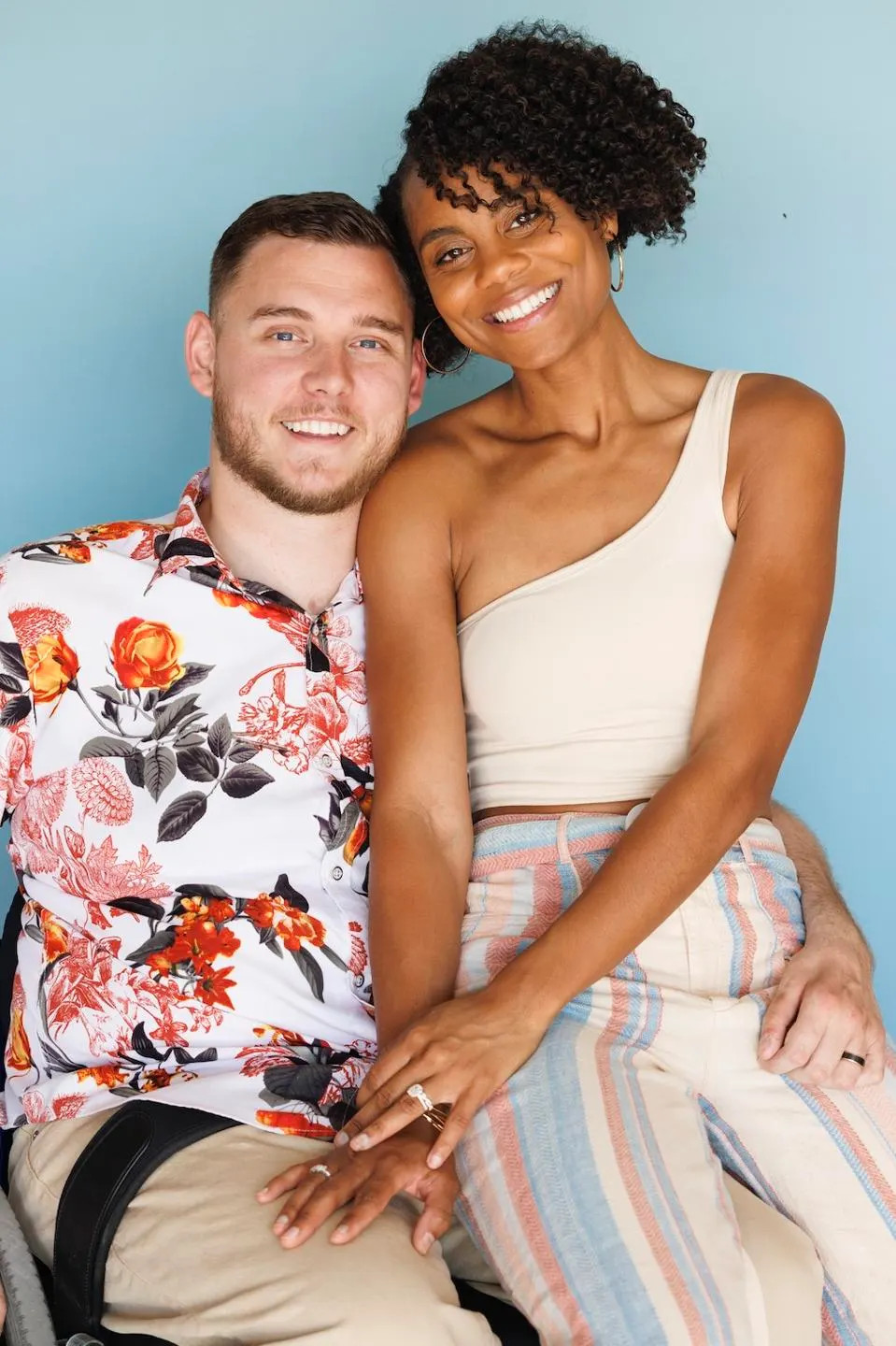 Cole and Charisma Sydnor are pictured in a photo from Forbes. Cole has light skin, short hair, a short brown beard, and a tropical short and is seated in his wheelchair. Charisma has brown skin, curly black hair, and a wedding ring visible on her left ring finger.