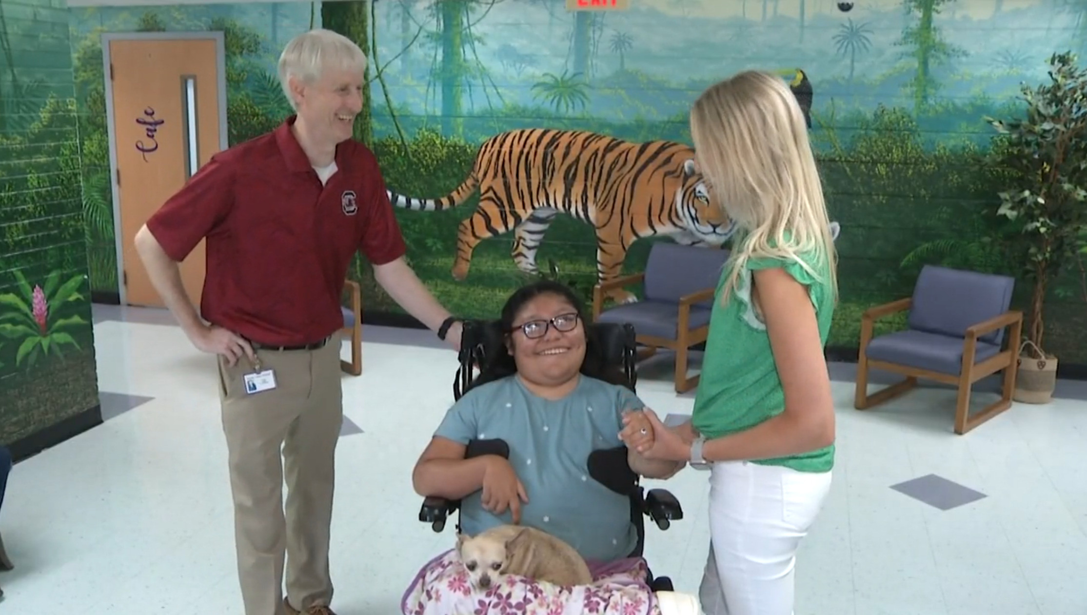 In a screenshot from news coverage, 11-year-old Cynthia Del Valle-Silvero is at her school with two staff members. She is sitting in her black power chair with her small service dog on her lap and has light brown skin, dark hair, and glasses.