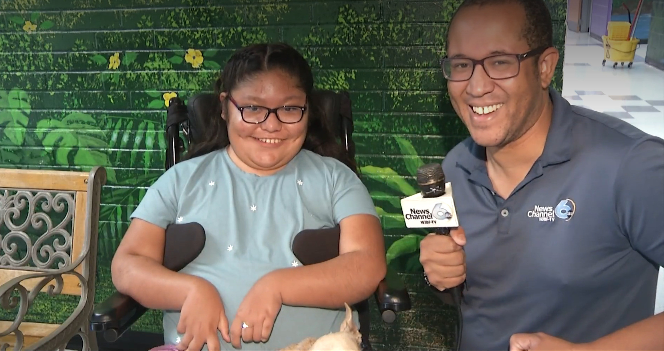 11-year-old Cynthia del Valle-Silvero is being interviewed by Brandon Dawson of WJBF Newschannel 6 ABC at her school. Cynthia has light brown skin, glasses, and dark hair and is seated in her power chair with a small service dog on her lap. Brandon has light brown skin, glasses, and a Newschannel 6 ABC polo shirt with an interview mic in his hand. Both are smiling.