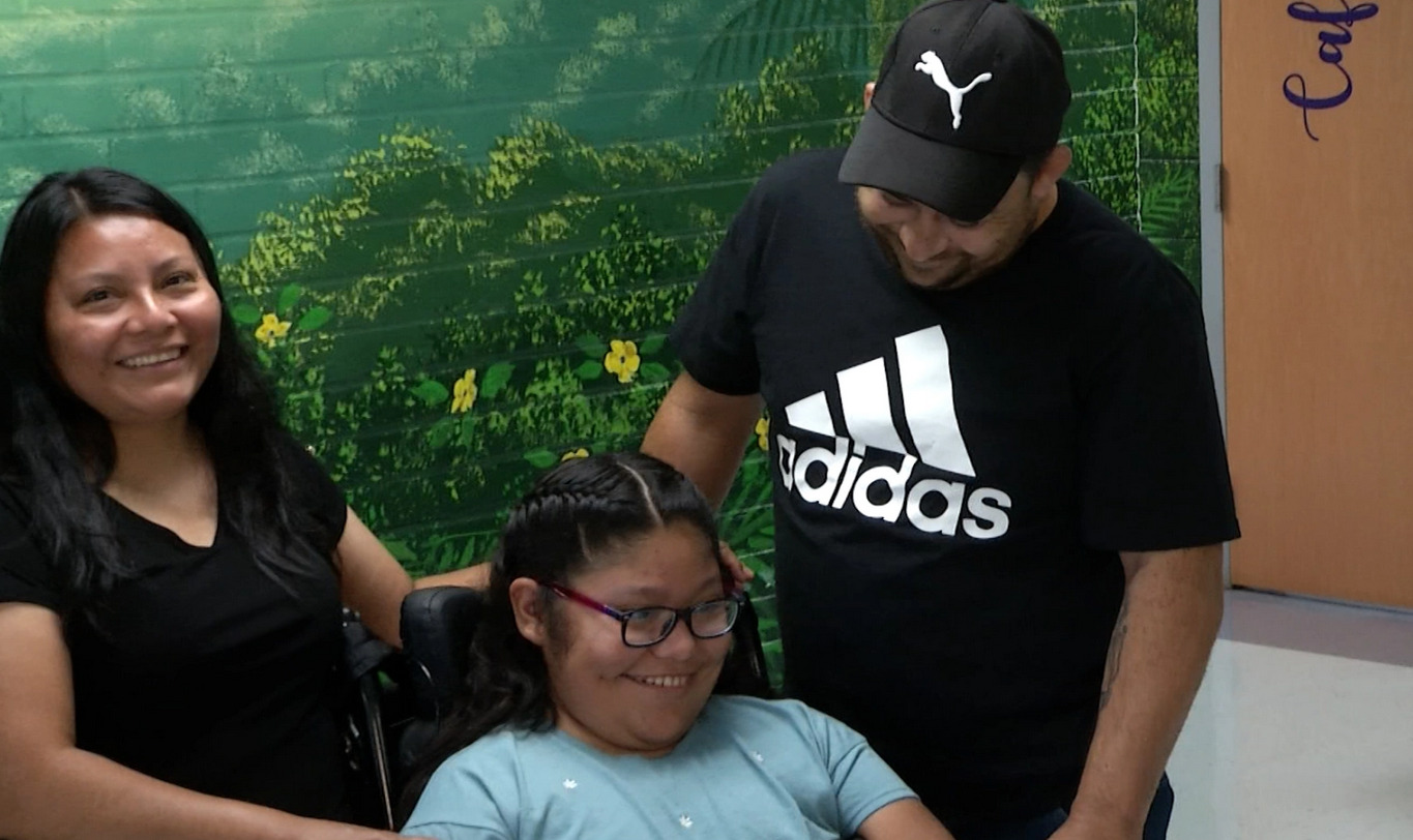 Cynthia Del Valle-Silvero smiles in a screenshot from news coverage filmed at her school. She is with her mom and dad - all three have light brown skin and dark hair. Cynthia has glasses and sits in her black power chair.