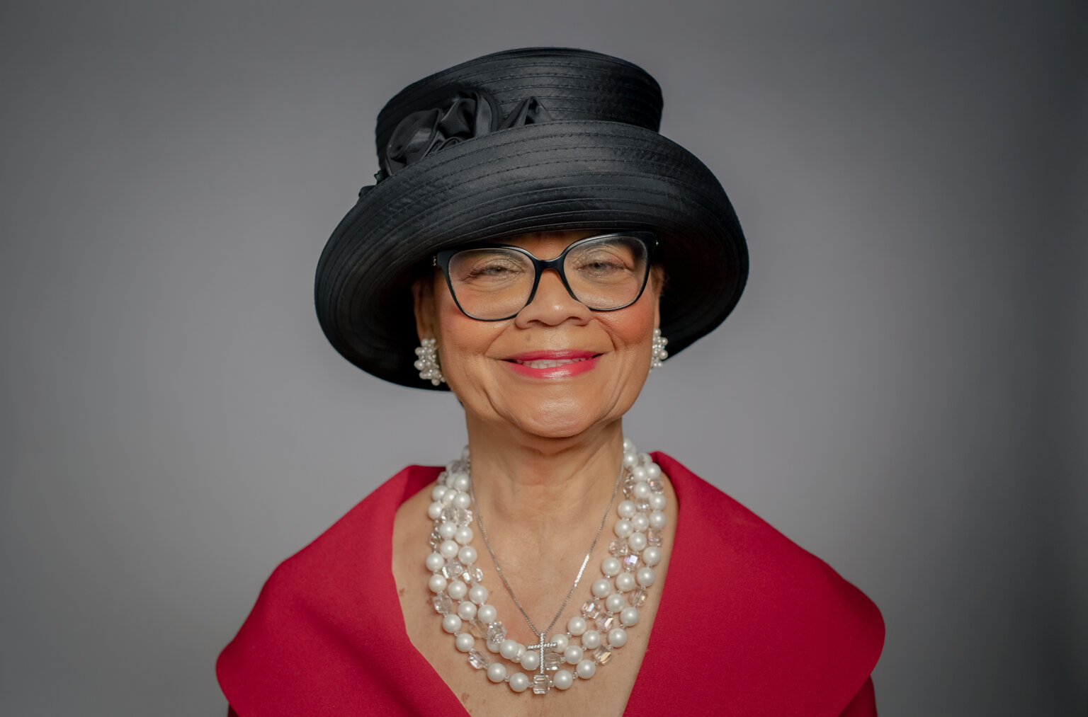 In a portrait for her Power Women 2023 profile, Rev. Dr. Lorina Marshall-Blake wears a red dress, pearl necklace, sparkling earrings, and a black decorative hat. She has brown skin, red lipstick, and black framed glasses.