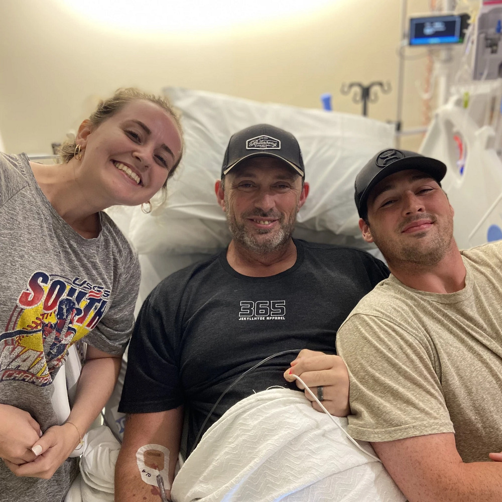 Heart transplant candidate Sean Hudson is pictured in a hospital bed with light skin, a short gray beard, and a ball cap. He is with two younger individuals, one male and one female.