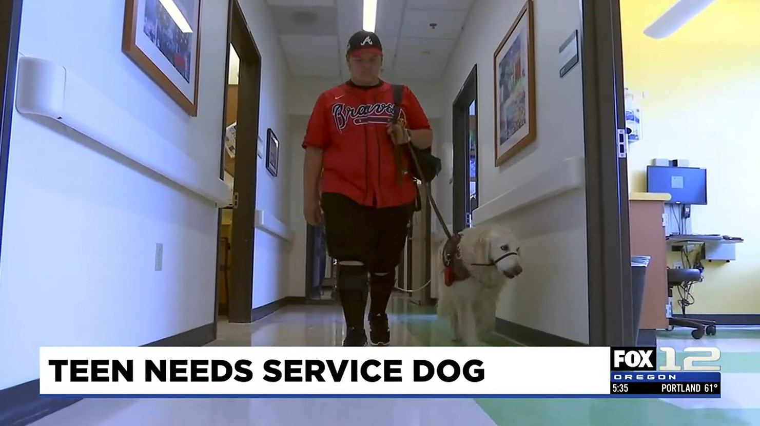 15-year-old Stryder Doescher wears a red Atlanta Braves jersey and a matching ball cap as he walks down a hospital corridor with his white retriever service dog, Keebler. A ticker from FOX 12 Oregon reads Teen Needs Service Dog.