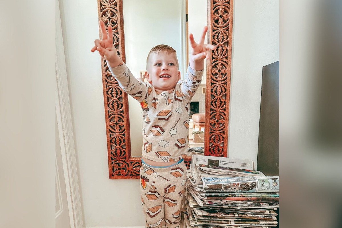 Three-year-old Zeke Clark is pictured pre-amputation giving peace signs at home. He has light skin, short brown hair, and a smores themed pajama set.