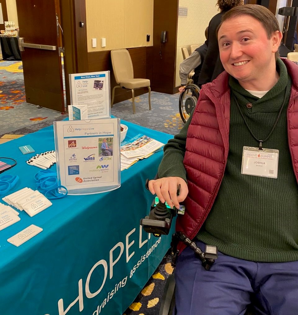 At a Reeve Summit event, Josh Basile smiles next to the Help Hope Live booth. Josh has light skin, brown eyes, and short brown hair and sits in his black power chair.