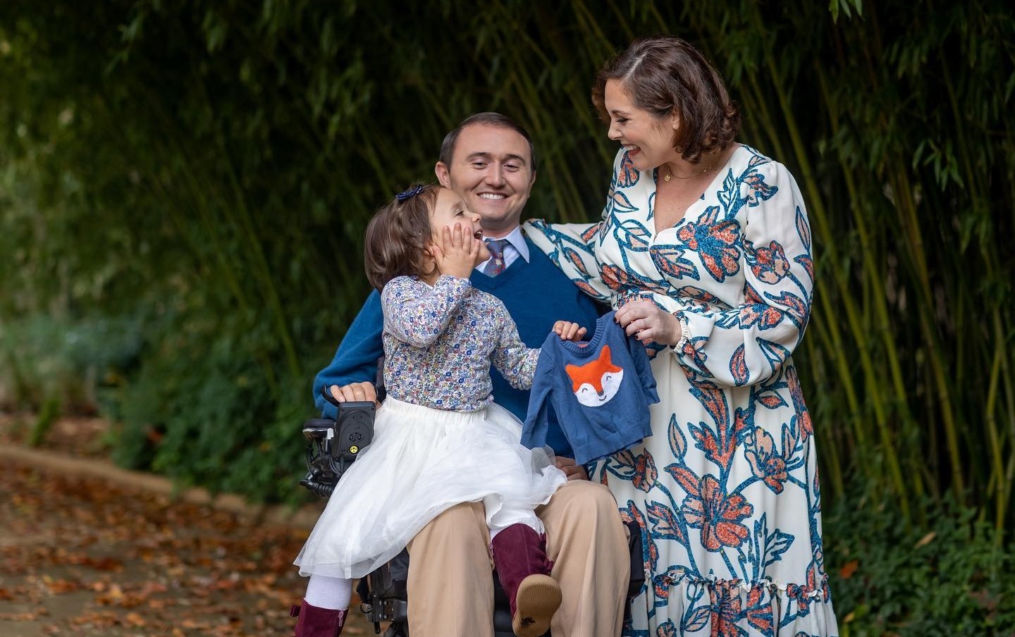 Josh and his wife are pictured with their young daughter in an outdoor photo shoot with a bamboo grove. Josh has light skin, short brown hair, and formal attire as he sits in his black power chair. His wife has light skin, curled brown hair, red lipstick, and a flower print white, orange, and blue long-sleeved dress. His daughter wears a white tutu skirt and holds up a blue child-sized sweater with a friendly fox face. All 3 are beaming.