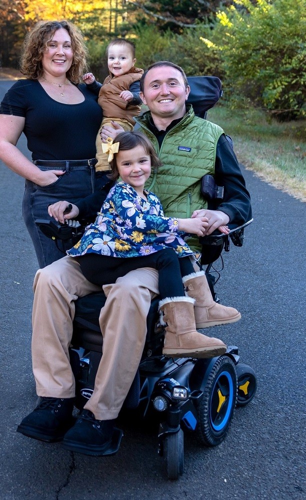 Josh Basile outdoors on a blacktop with his family. Josh has light skin, short brown hair, and a green vest and khakis. He is seated in his black power chair with his young daughter on his lap in a flowery top and fuzzy boots. His wife has light skin and short curly hair and holds his toddler son in her left arm as she stands beside Josh's power chair.