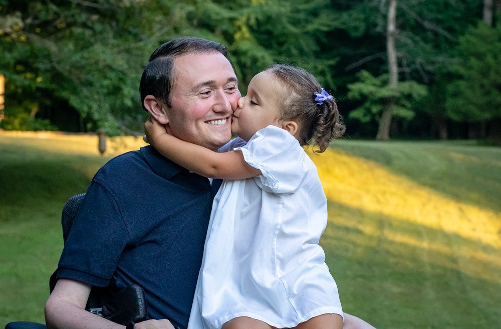 In an outdoor photo on a grassy expanse, Josh gets a big kiss on the cheek from a little girl with light skin and curly brown hair in a white dress as she sits on his lap. Josh has light skin, short dark hair, and a blue polo and sits in his black power chair.