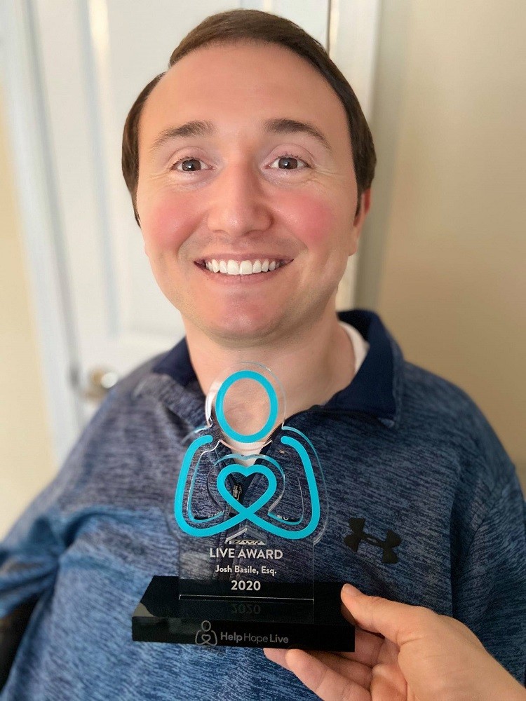 Seated in his wheelchair, Josh smiles with light skin, rosy cheeks, brown eyes, and short brown hair a someone holds up his Live Award from Help Hope Live in front of his chest. The award has a teal Help Hope Live pledge figure logo and the inscription Live Award: Josh Basile, Esq. 2020.