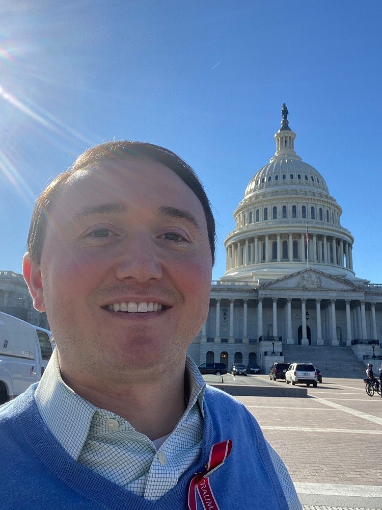 Josh Basile takes a selfie in front of the capital building. He has light skin, short brown hair, brown eyes, and a collared shirt and blue vest. He wears a ribbon in red trimmed in white that reads TRAUMA.